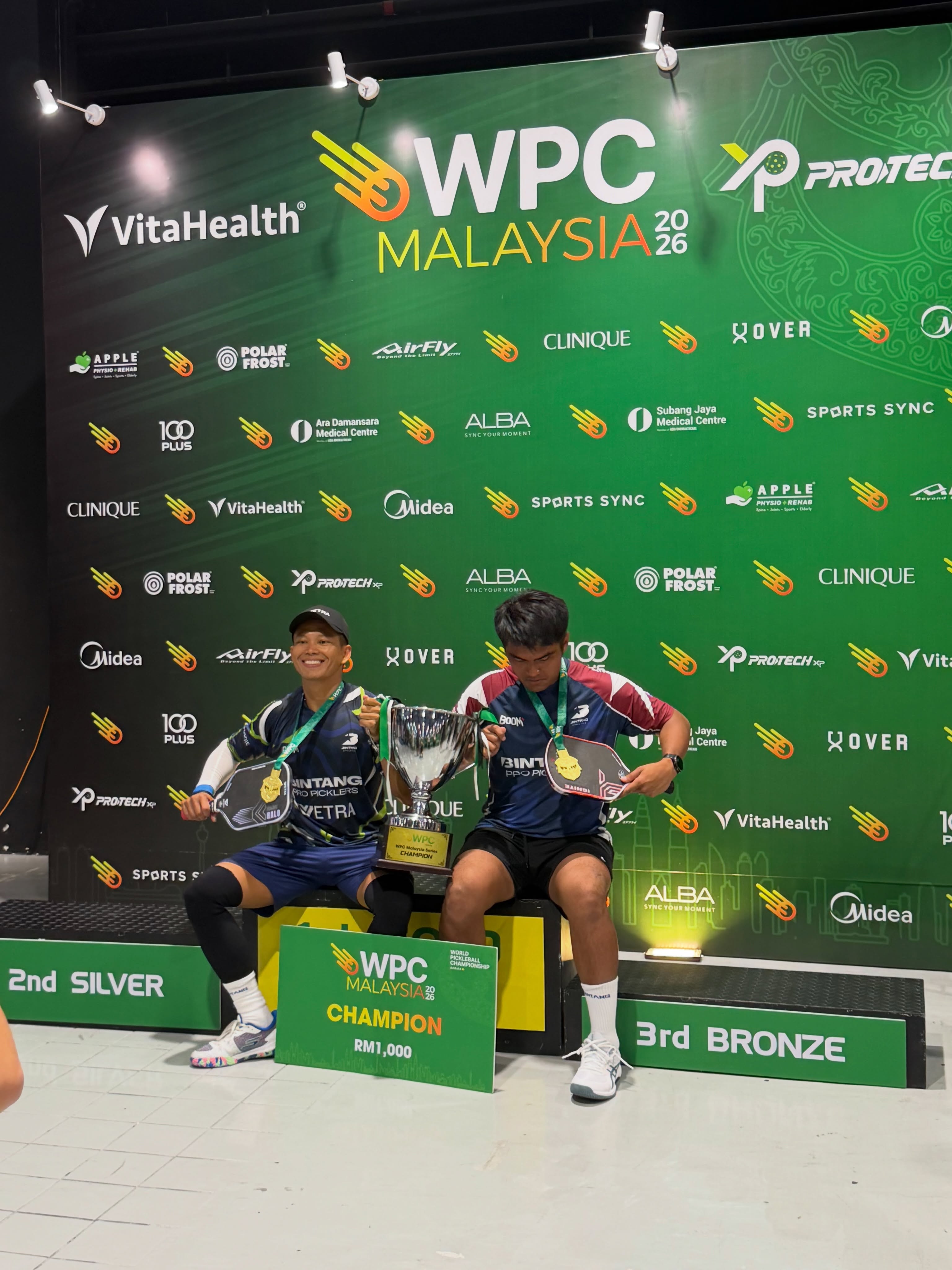 Ashaari Zainal sits with his Double 19+ 4.5 Advanced gold-medal partner, holding the champion trophy on the podium.