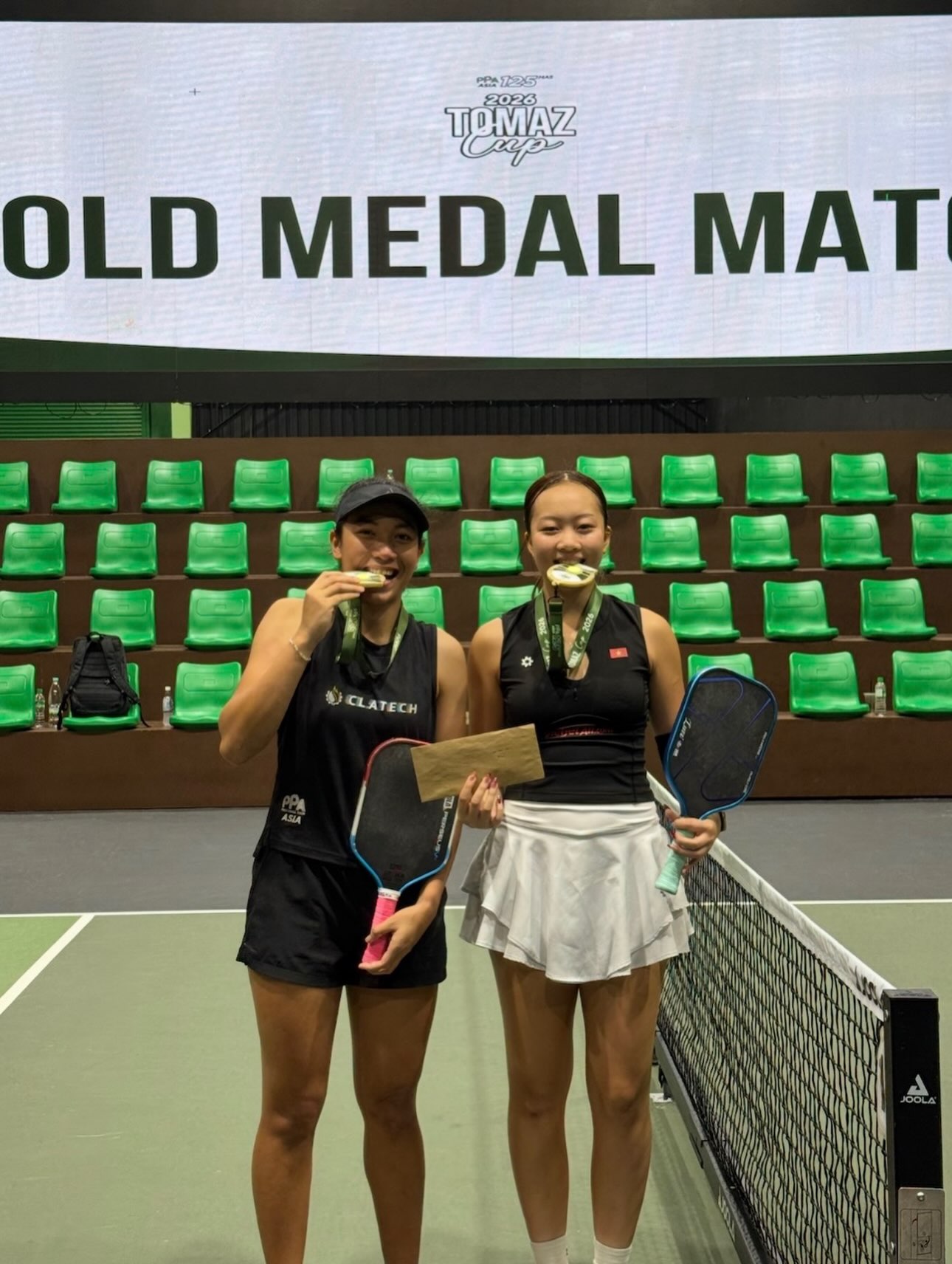 Tya Karina (left) and Sophia Tran Phuong Anh (right) bite their gold medals in front of the Tomaz Cup Gold Medal Match screen.