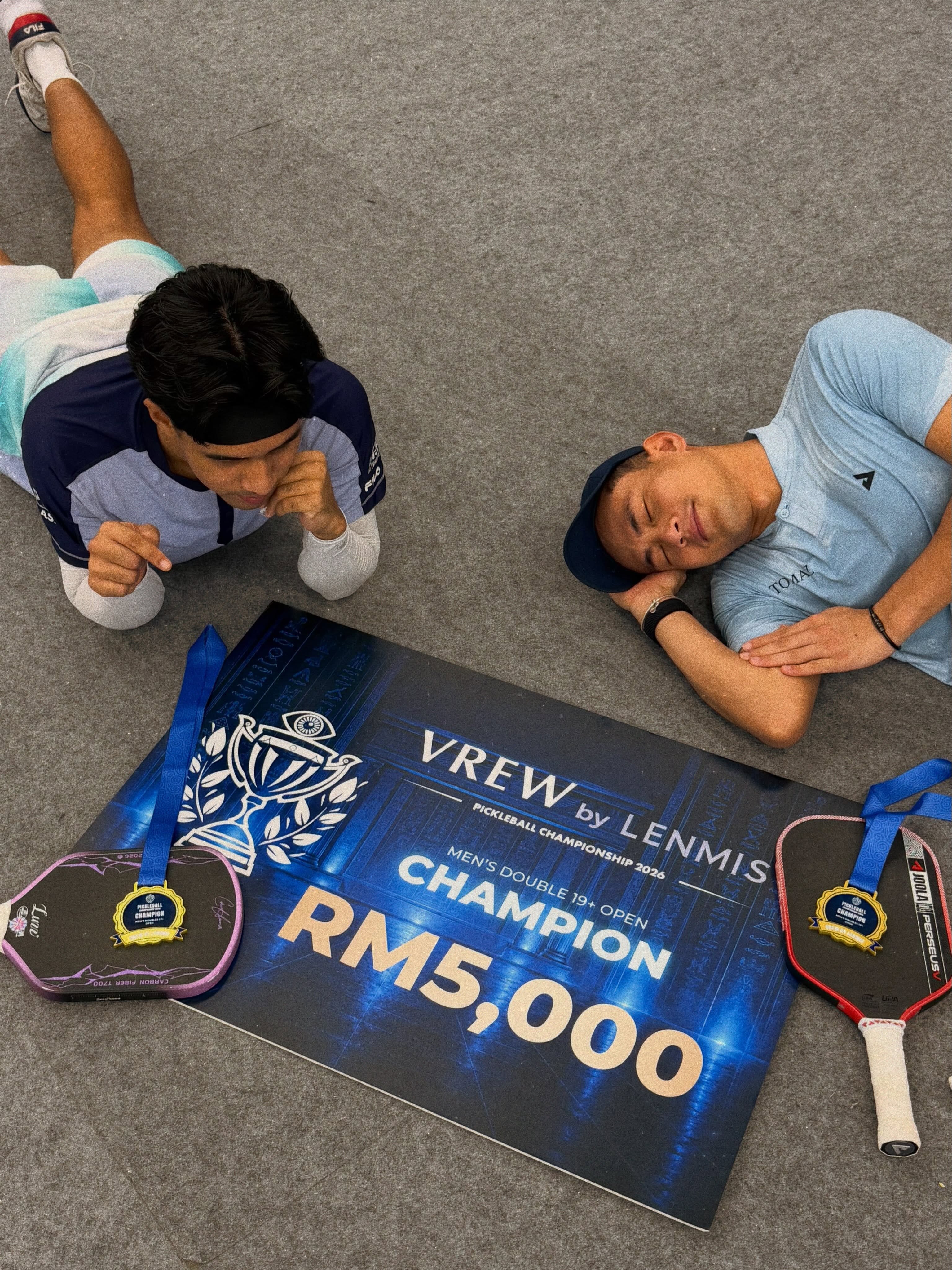Syed Uzair and Aaiman Hamdan lying on the floor next to their medals, paddles and a champion board reading RM5,000