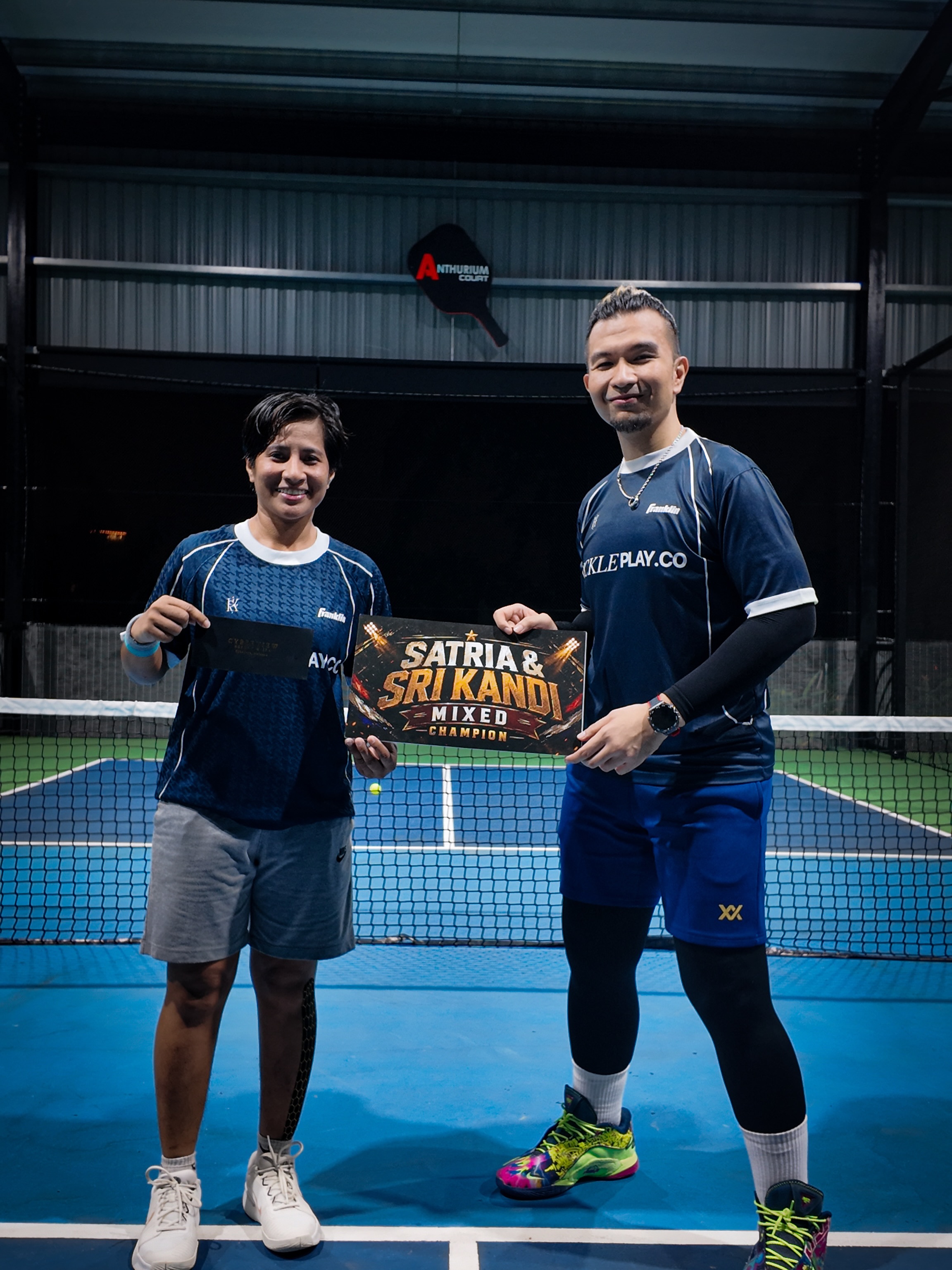 Two PicklePlayCo players holding a Satria and Sri Kandi Mixed Champion board on court