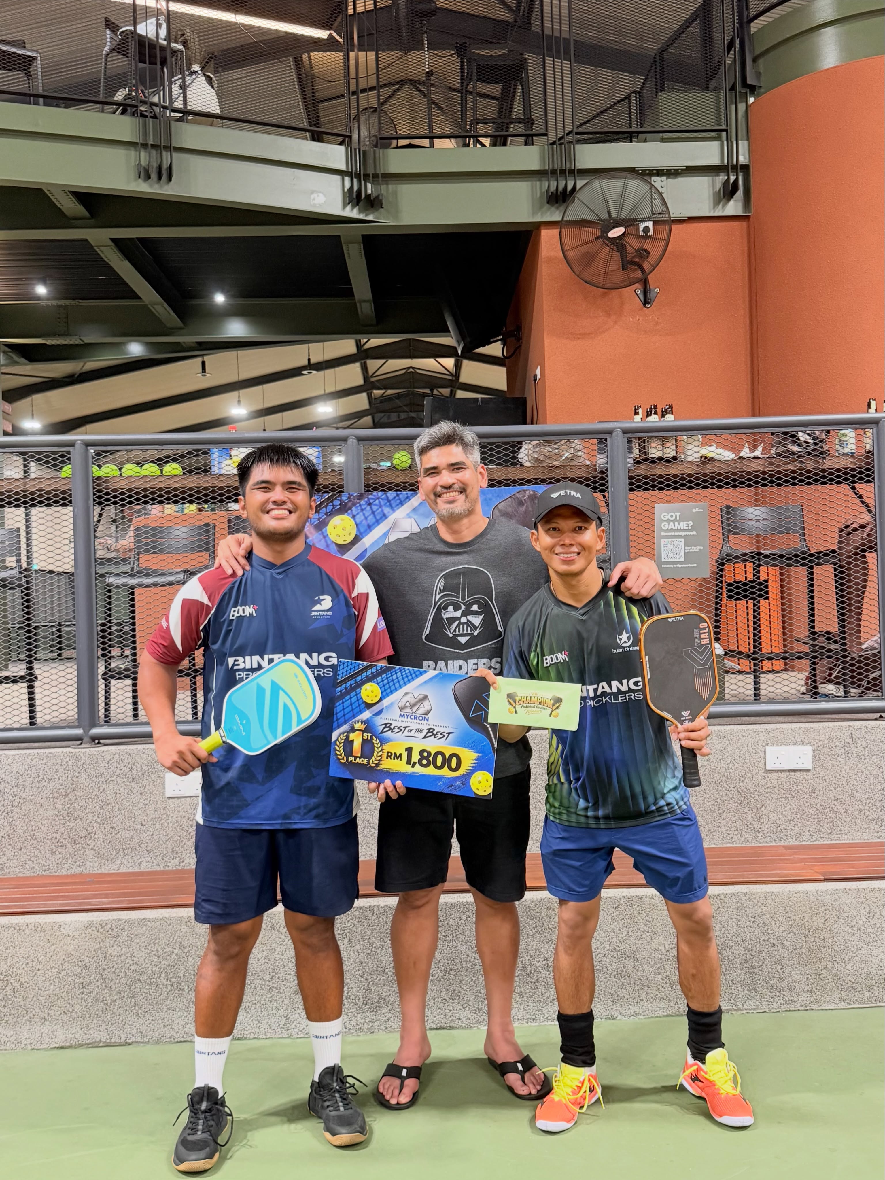 Two players in Bintang Pro Picklers jerseys holding the Mycron Invitational Best of the Best 1st place RM1,800 board with a third man between them