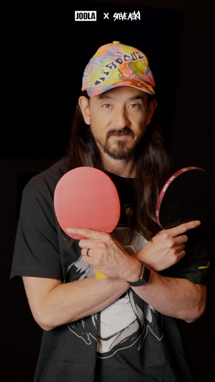 Steve Aoki holding two table tennis rackets crossed in front of his chest, with a JOOLA x Steve Aoki logo above