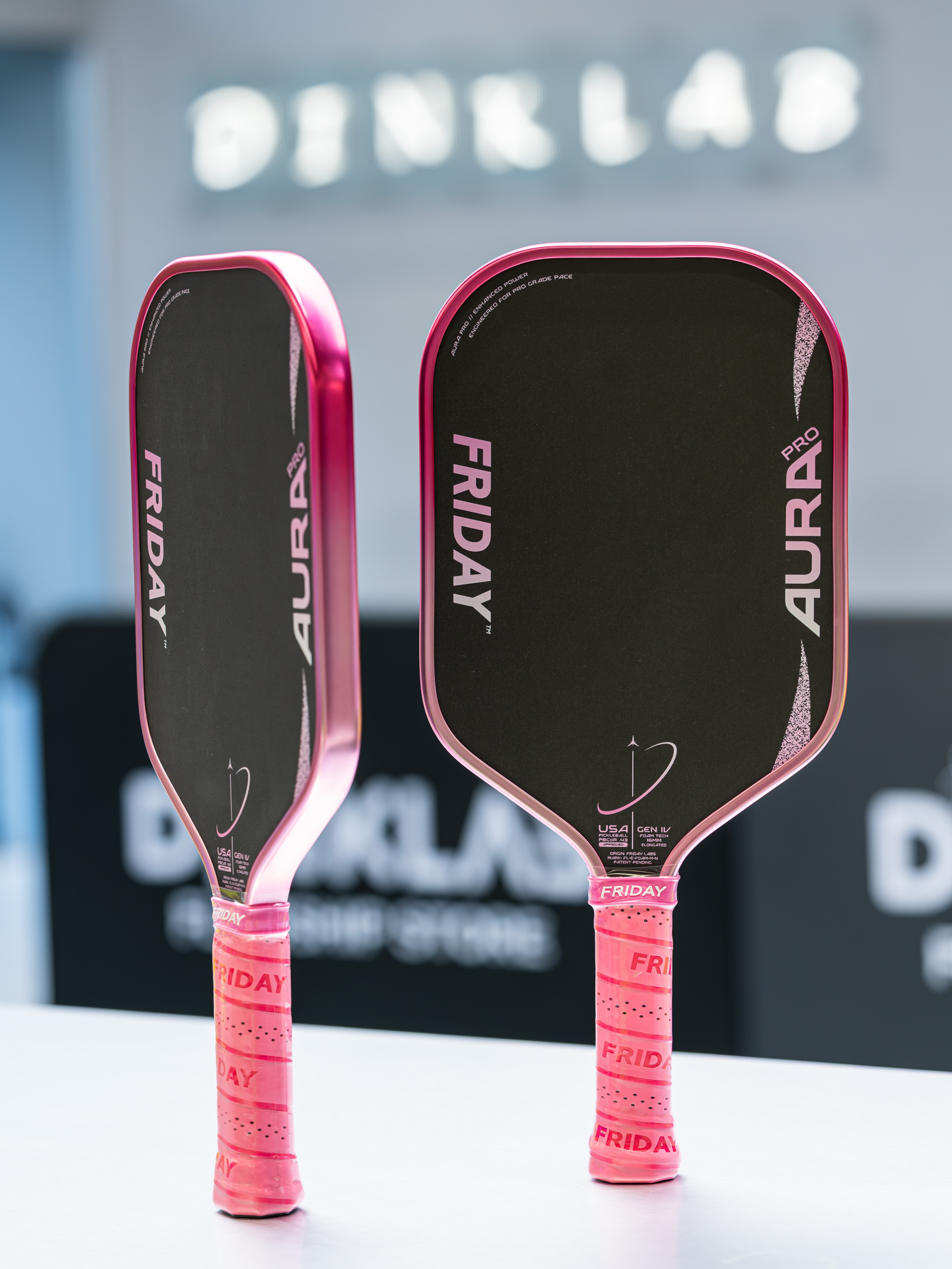 Two Friday Aura Pro Elongated Metallic Pink Prism paddles stand upright with pink grips in front of a Dinklab sign.