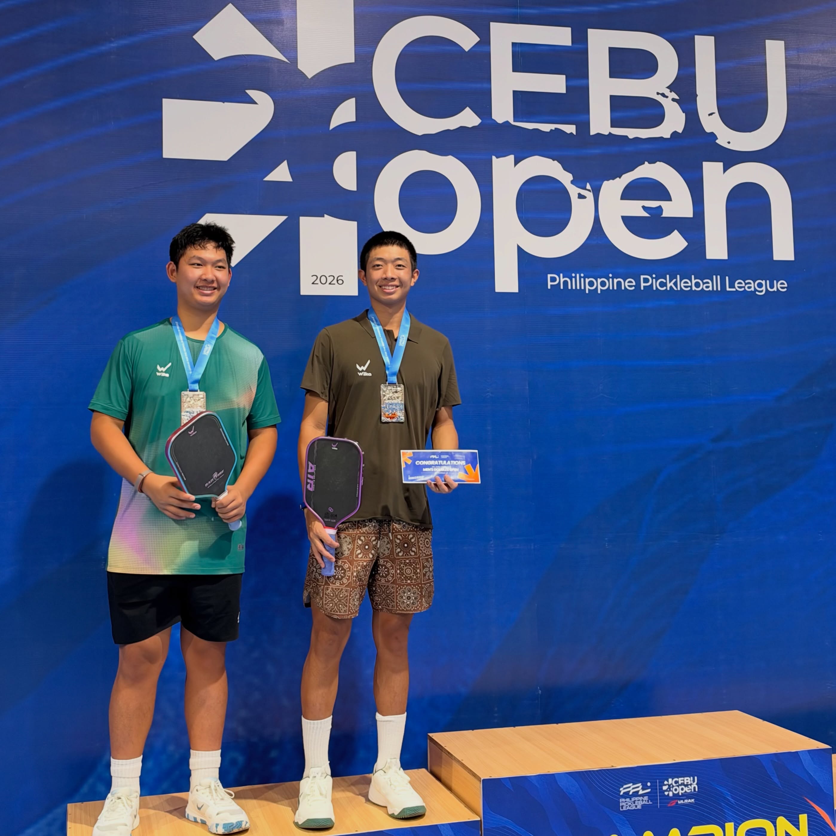 Two Wika-kitted players holding silver medals and a Men's Doubles Open certificate at the Cebu Open podium