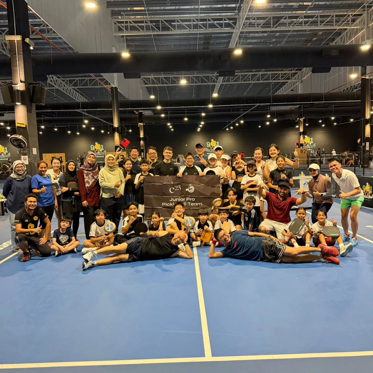 A group photo in front of an e23 Junior Pro Pickleball Team banner, with pans and cutting boards in hand