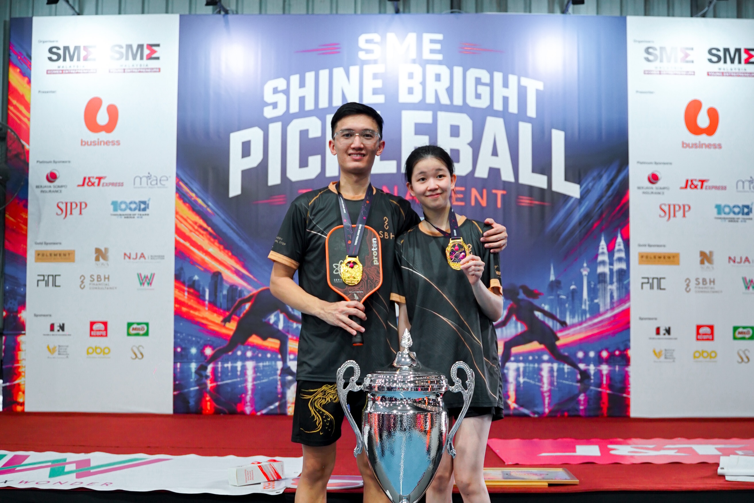 Two players in SBH shirts with gold medals standing beside the champion's trophy