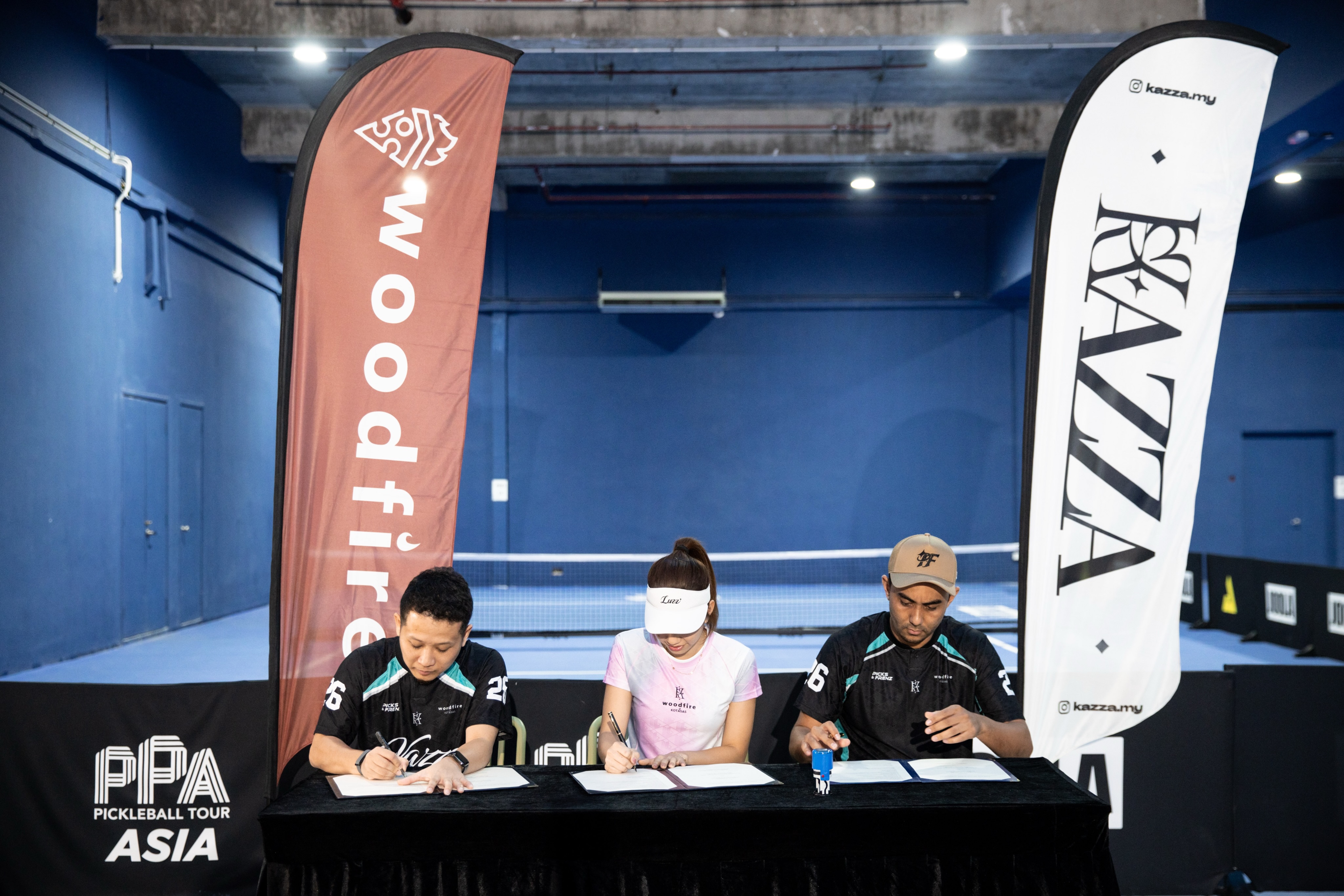 Three people signing documents at a table between Woodfire and Kazza banners