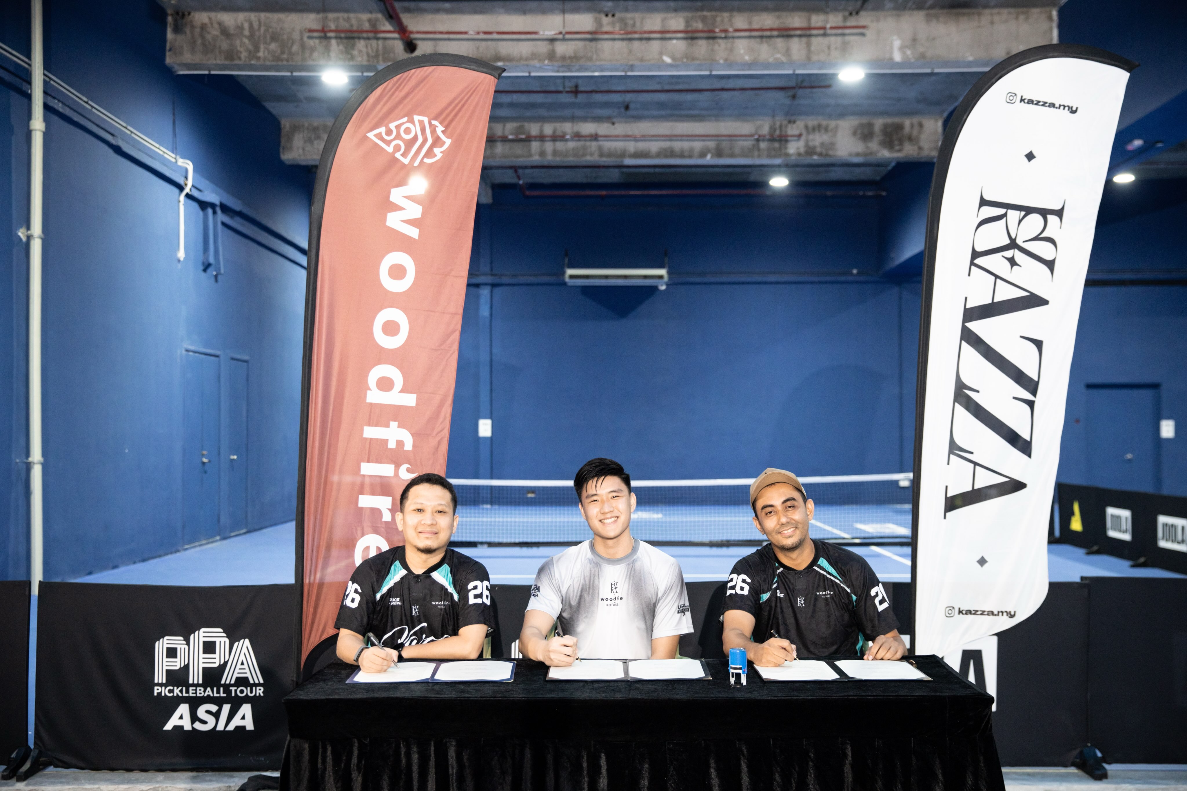 Three people seated at a table signing documents between a Woodfire banner and a Kazza banner, with PPA Pickleball Tour Asia branding behind