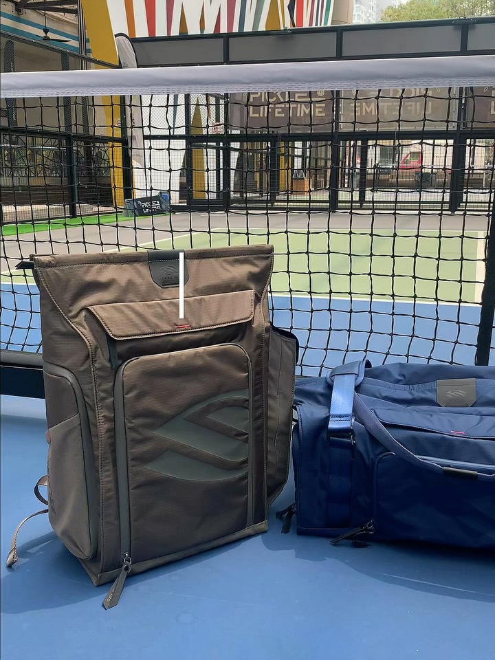 A Selkirk backpack and a navy Selkirk duffle bag courtside in front of a pickleball net
