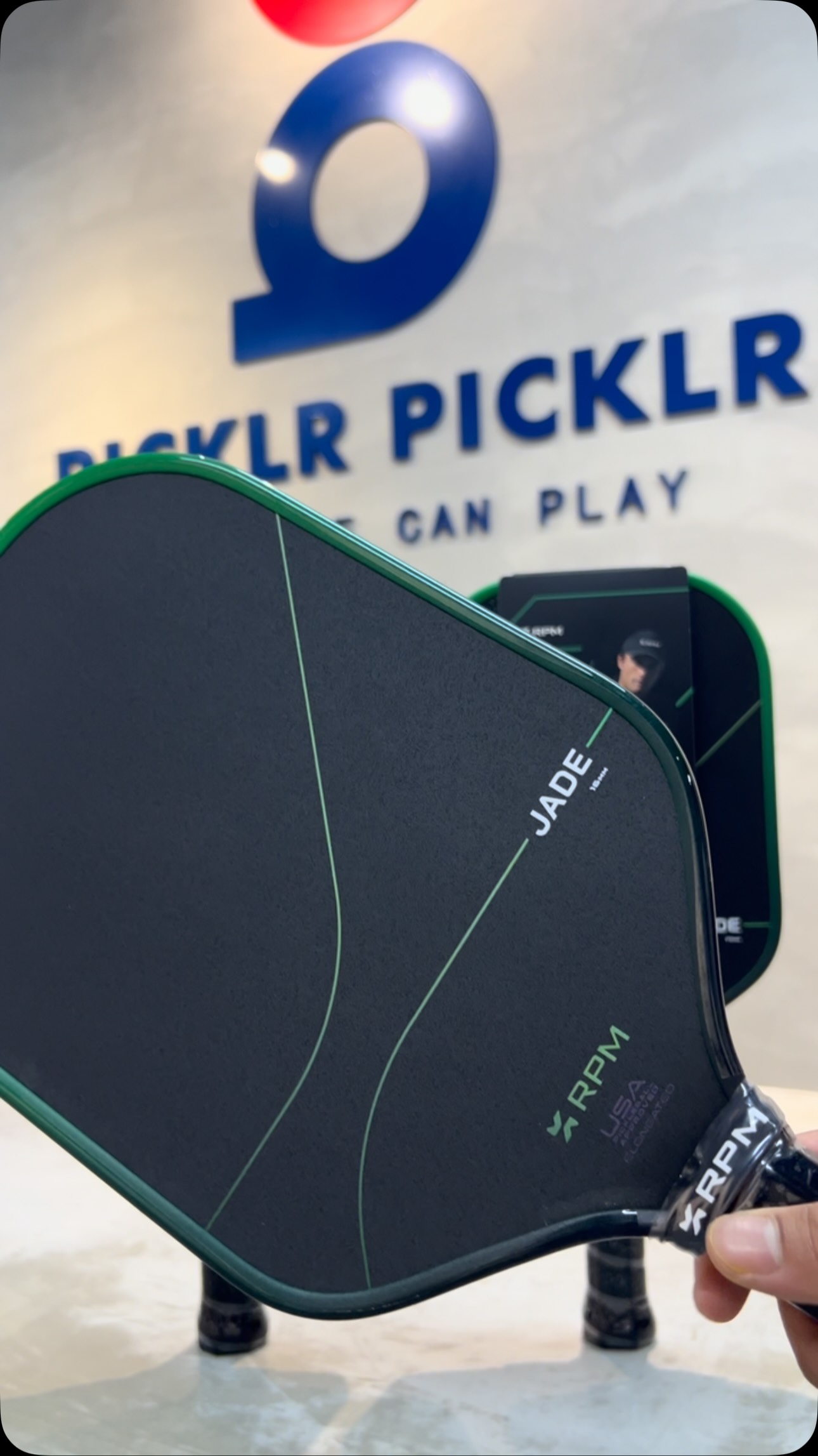 RPM Jade paddle face held in front of the Picklr Picklr wall signage