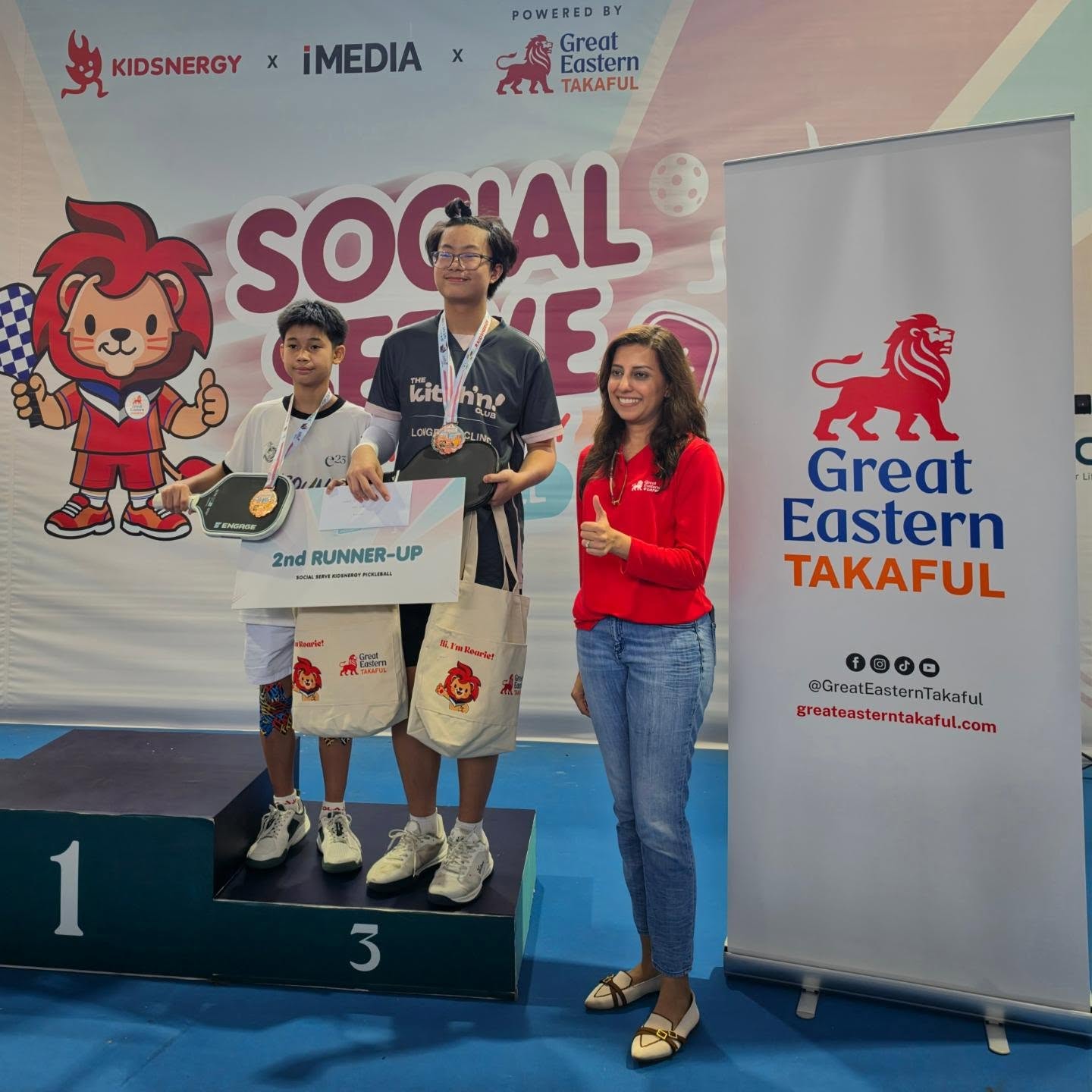 Two juniors with medals on the podium beside a 2nd Runner-Up board at Social Serve Kidsnergy Pickleball