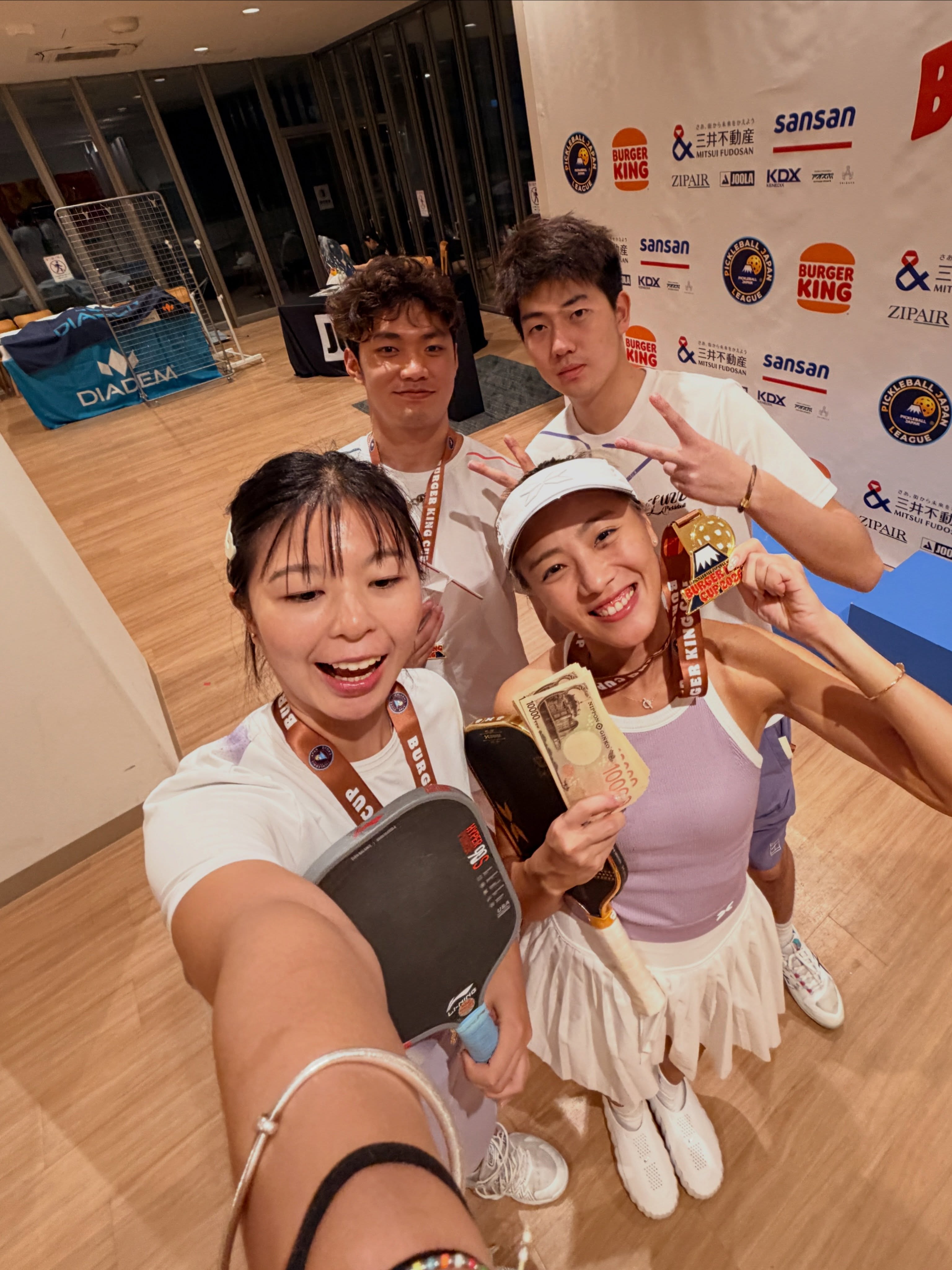 Four players in a selfie with medals, paddles and yen notes in front of the Burger King Cup wall