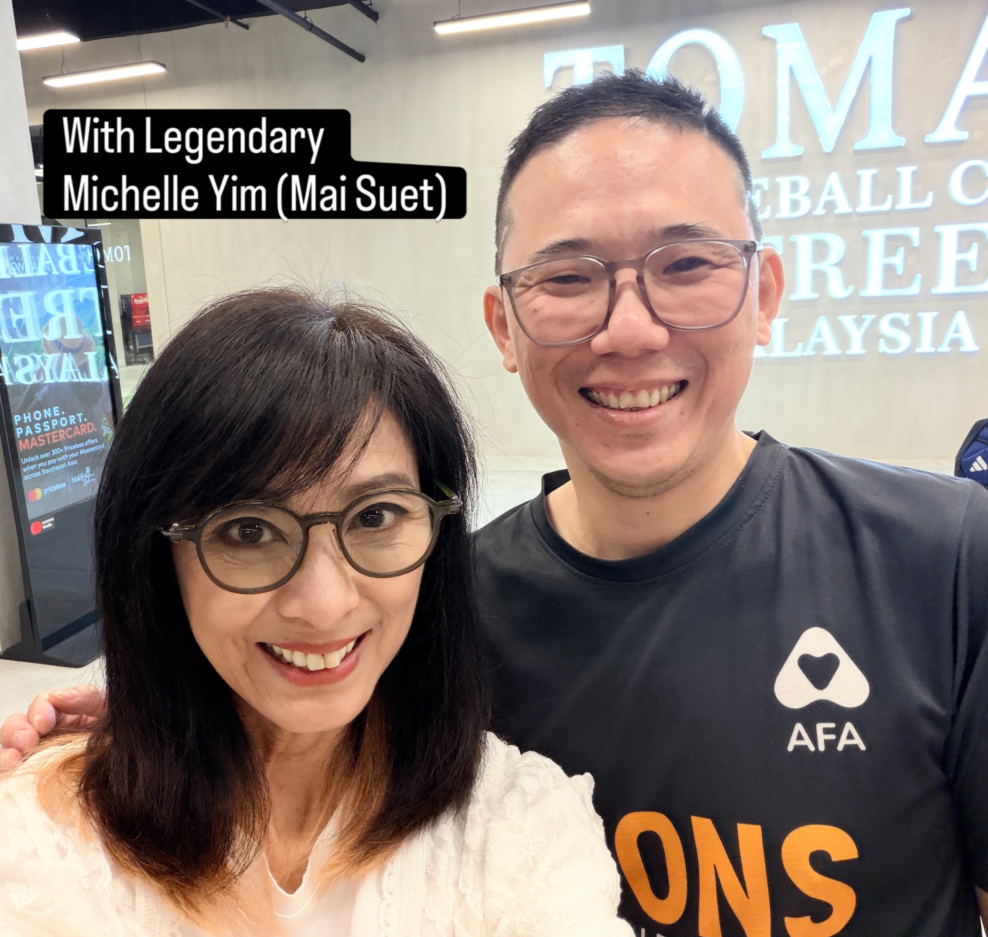 A man in a black shirt beside a woman in glasses, labelled With Legendary Michelle Yim (Mai Suet), in front of a partly visible Tomaz sign.