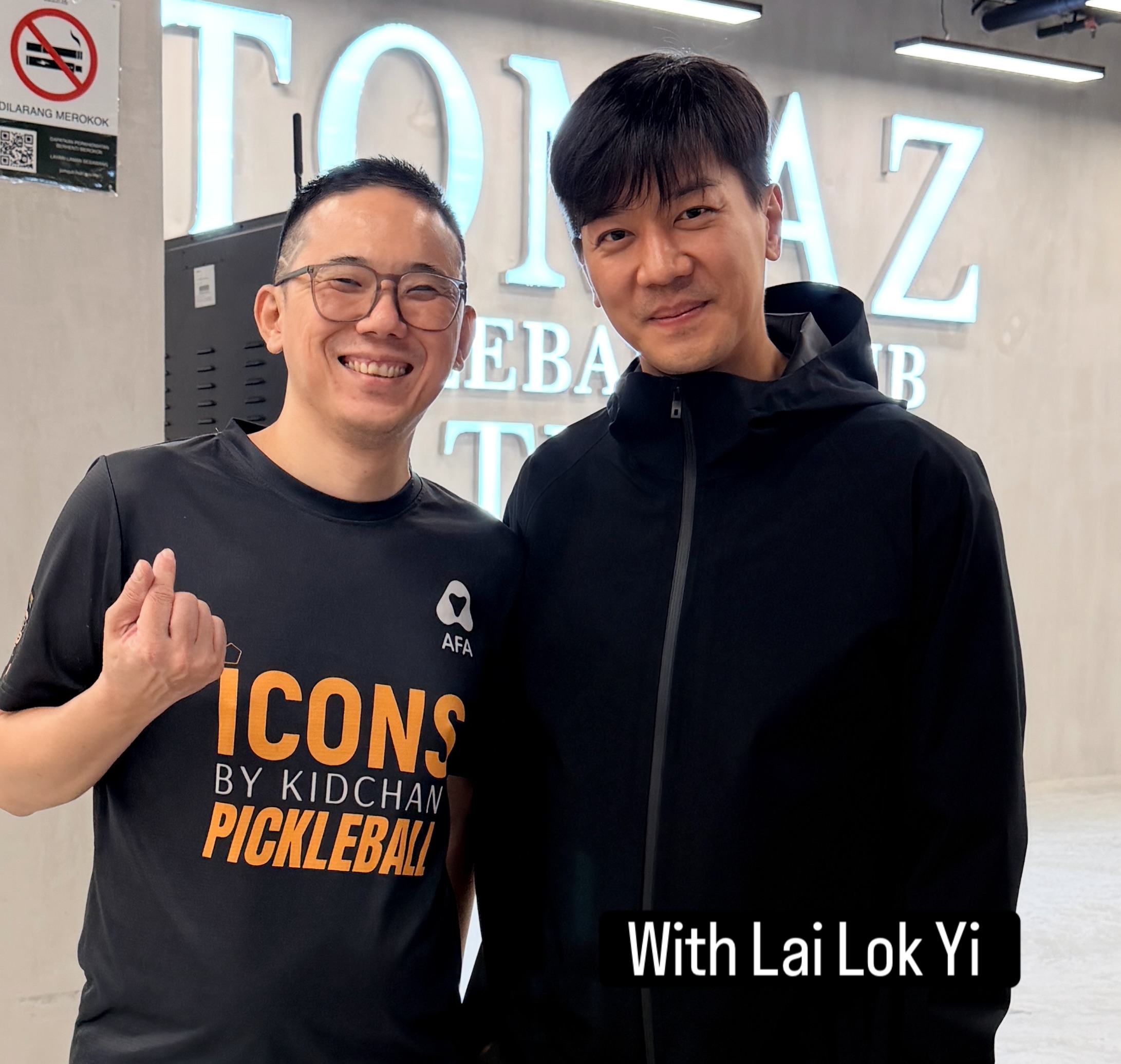 A man in a black shirt beside a man in a black jacket in front of a partly visible Tomaz sign, labelled With Lai Lok Yi.