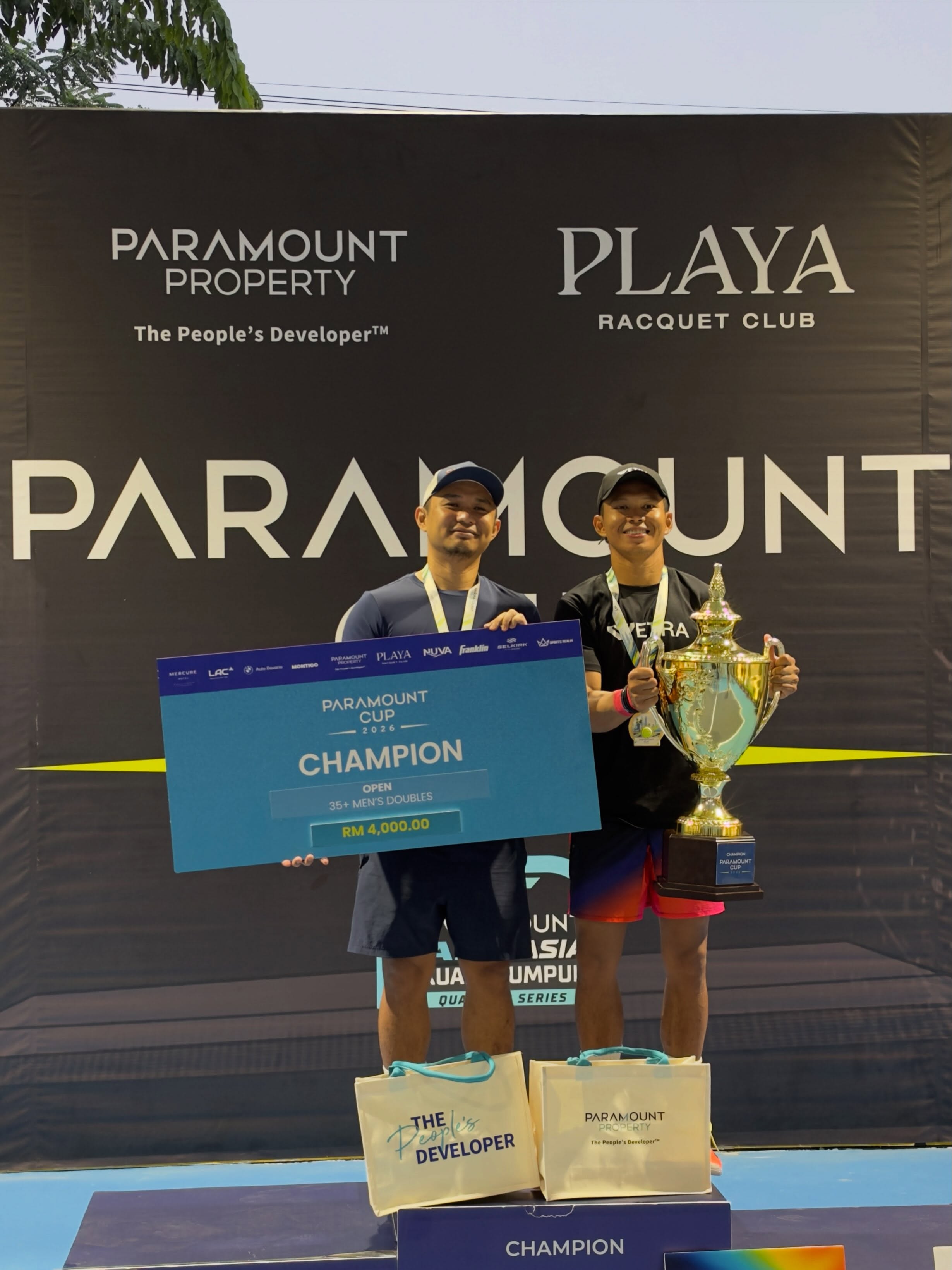 Two men on the podium holding a trophy beside a Paramount Cup champion placard reading Open 35+ Men's Doubles, RM4,000