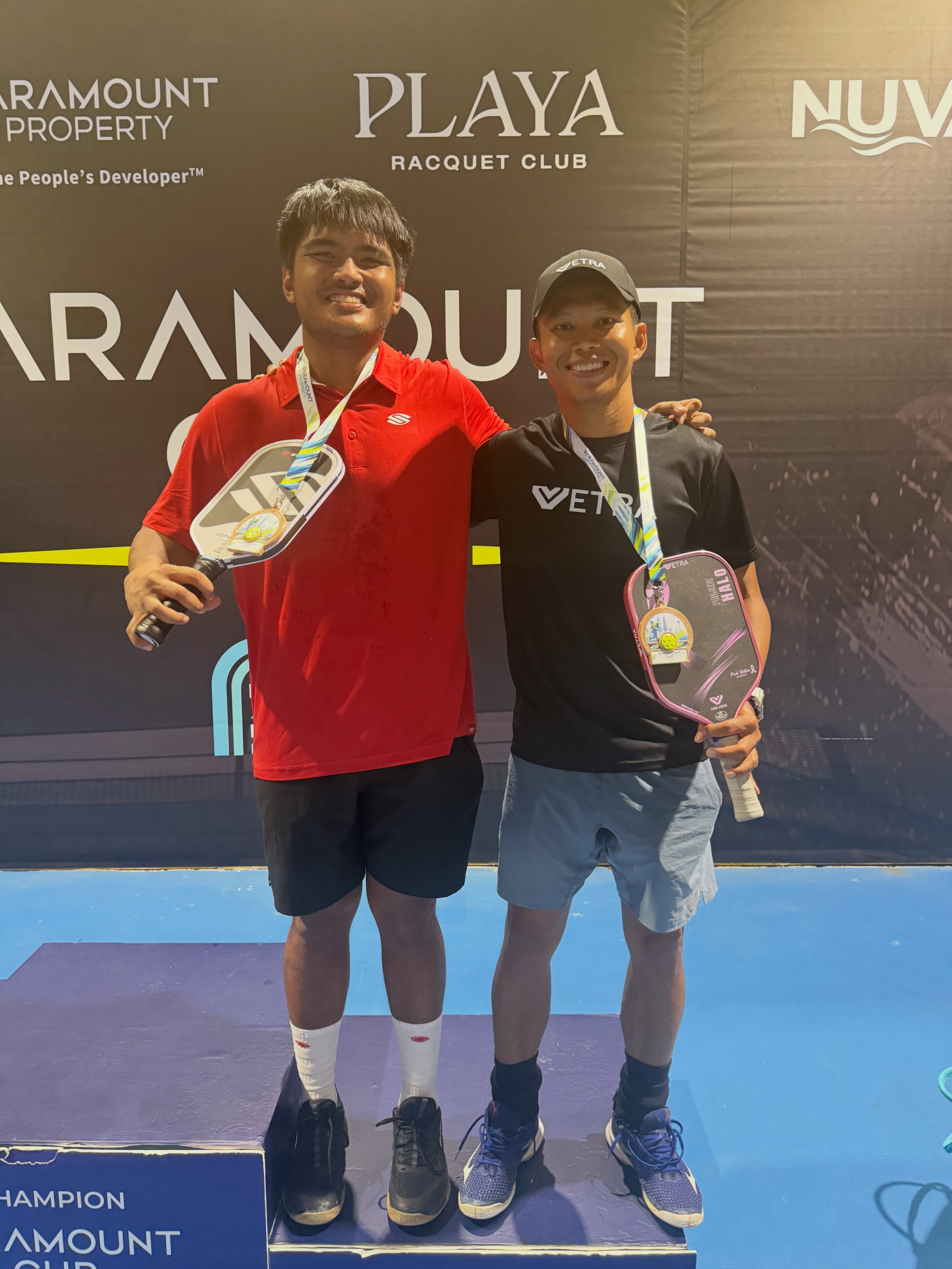 Two men posing on a Paramount Cup podium, one in red holding a paddle and one in a Vetra shirt