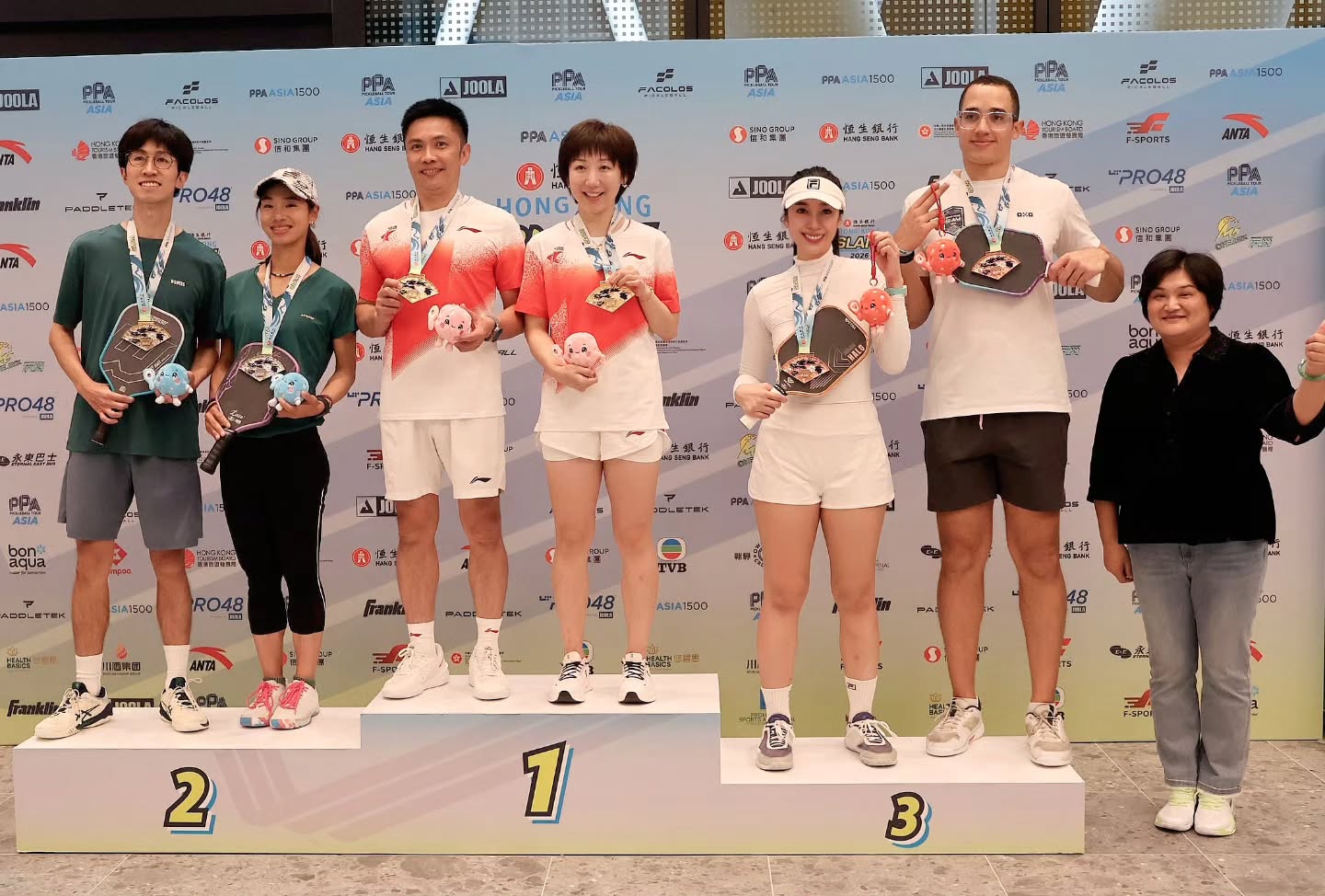 Regine Hoe, in a white outfit and Fila visor, stands with her partner on the third step of the podium at the PPA Hong Kong Slam 2026, both holding bronze medals
